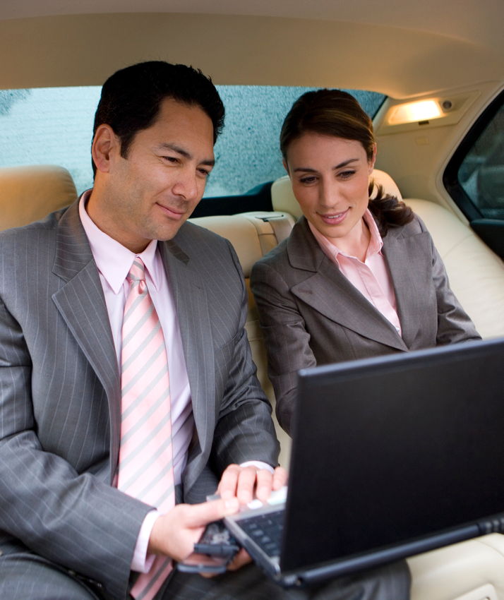 A businessman and a businesswoman are sitting in the back seat of a chauffeur-driven limousine, concentrating on a laptop computer.